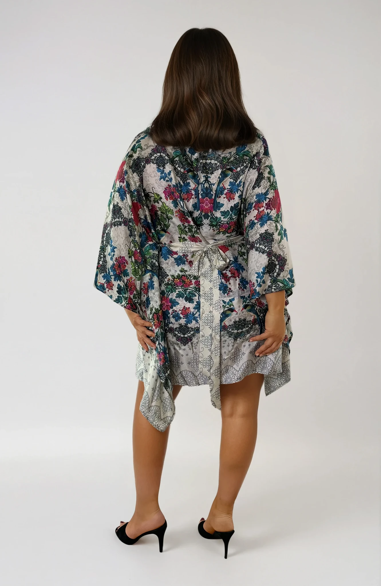 Back view of the ZALA silk kaftan dress in Secret Garden, featuring an ivory ground adorned with painterly florals in soft teal, berry, rose, and sage tones. Inspired by antique botanical textiles, the intricate pattern evokes timeless romance, artistry, and effortless elegance. The removable sash is tied at the back to softly define the waist, while flowing draped sleeves and an asymmetrical hem create graceful movement from every angle.